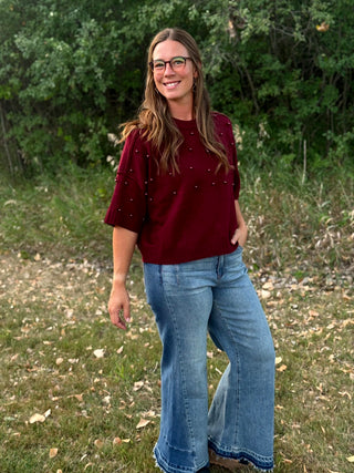 A woman wearing a burgundy short-sleeve knit top with small bead embellishments and light blue flare jeans, standing outdoors in a grassy area with trees in the background.