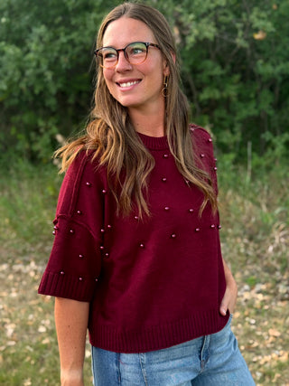 A woman wearing a burgundy knit sweater with silver bead accents on the sleeves and chest, paired with blue jeans, standing outdoors in front of a blurred green background.