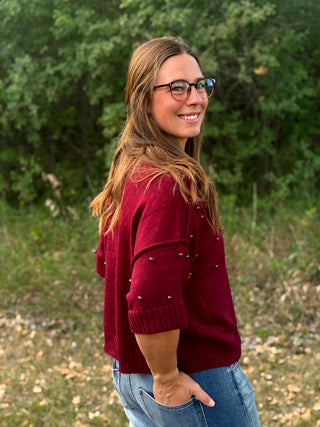A woman with glasses and long light brown hair is shown from the back and side, wearing a maroon knit sweater with pearl embellishments on the sleeves and blue jeans, standing outdoors in front of a green wooded background.