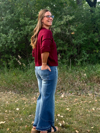 A woman wearing a burgundy knit short-sleeve top with decorative buttons and blue jeans, standing outdoors in a grassy area with trees in the background.
