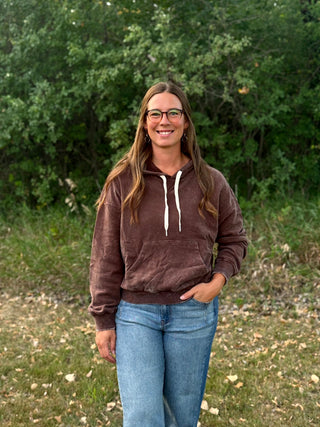 A woman with long light brown hair and glasses stands outdoors in front of a green bush, wearing a brown ribbed knit hoodie with white drawstrings and blue jeans.