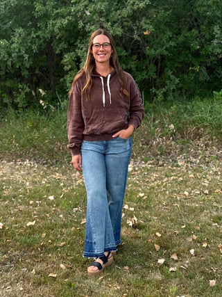 A woman wearing a brown pullover hoodie with white drawstrings, blue jeans, and black sandals, standing outdoors on a grassy lawn with trees in the background.