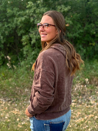 A woman with long brown hair and glasses, wearing a brown corduroy hoodie and blue jeans, standing outdoors in front of a green wooded background.