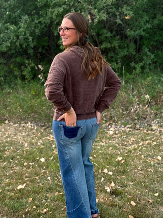 A woman wearing a brown corduroy hoodie and blue jeans with dark blue patch pockets, standing outdoors in a grassy field with trees in the background.
