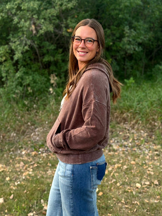 A woman with long brown hair and glasses stands outdoors in a wooded area, wearing a brown corduroy hoodie and blue jeans.
