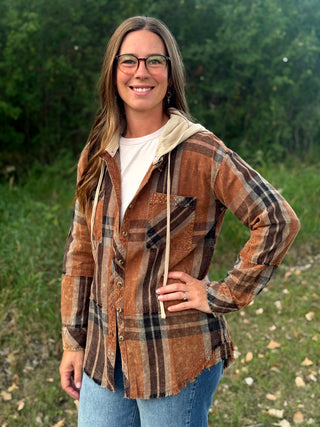 A woman wearing a brown and tan plaid hooded button-up shirt with a beige fleece-lined hood, layered over a white t-shirt and blue jeans, standing outdoors in front of a green forest background.