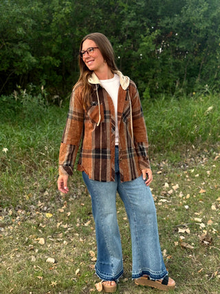 A woman wearing a brown and black plaid hooded flannel shirt over a white t-shirt, blue wide-leg jeans, and brown sandals, standing outdoors in a grassy field with trees in the background.