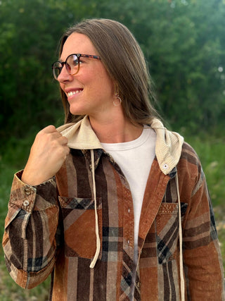  woman wearing a brown and tan plaid hooded button-up shirt with a beige fleece-lined hood, layered over a white t-shirt and blue jeans, standing outdoors in front of a green forest background.