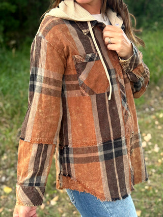 A woman wearing a rust-orange and brown plaid patchwork hoodie jacket over a cream hoodie, paired with blue jeans, photographed outdoors in a grassy setting.