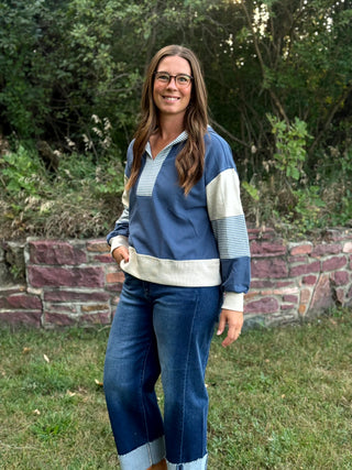 A woman wearing a blue and cream color-blocked long-sleeve knit top and blue jeans, standing outdoors in front of a stone wall and green foliage.