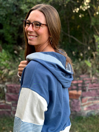 A woman wearing a blue, white, and light blue striped color-block hoodie, shown from a three-quarter back view in an outdoor garden setting.