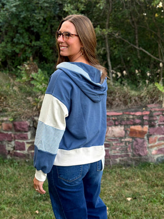 A woman wearing a blue and white color-block hoodie with striped details on the sleeves, standing outdoors in front of a stone wall and greenery.
