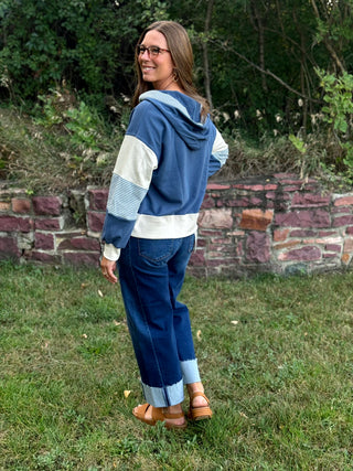 A woman wearing a blue and white color-blocked hoodie, blue jeans, and brown sandals stands outdoors on a grassy lawn in front of a stone wall and trees.