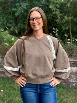 A woman wearing a taupe brown crewneck sweatshirt with white lace trim on the shoulders and cuffs, paired with blue jeans, standing outdoors in front of a green garden background.