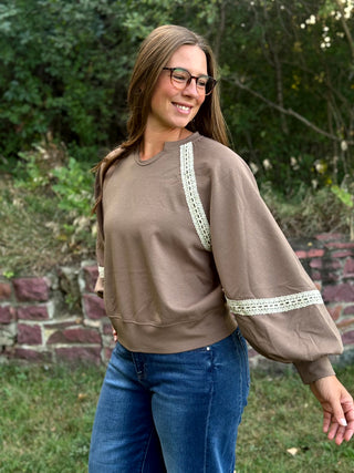 A woman wearing a taupe brown crewneck sweatshirt with white lace trim on the shoulders and sleeves, paired with blue jeans, standing outdoors in front of a stone wall and green foliage.