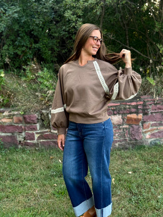 A woman wearing a brown long-sleeve top with white lace trim on the sleeves and blue jeans, standing outdoors in front of a stone wall and green foliage.