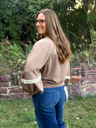 A woman wearing a taupe brown long-sleeve top with cream lace trim on the cuffs and blue jeans, standing outdoors in front of a stone wall and green foliage.