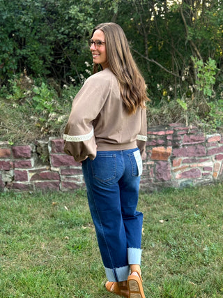 A woman wearing a beige bomber-style top with white lace trim on the sleeves, blue jeans with light blue cuffs, and tan shoes, standing outdoors on grass in front of a brick wall and trees.