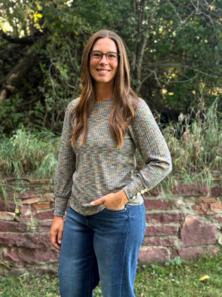 A woman with long brown hair and glasses stands outdoors in front of a stone wall and greenery, wearing a long-sleeve multicolor knit sweater and blue jeans.