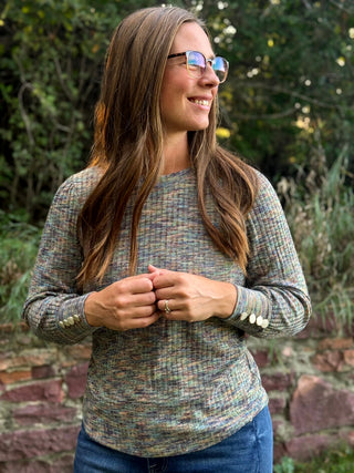 A woman wearing a long-sleeve knit top with a multicolor heathered pattern in muted green, blue, and beige tones, standing outdoors in front of a stone wall and greenery.