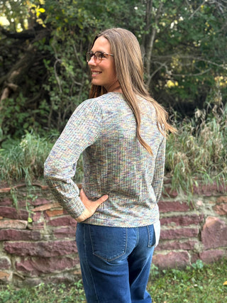 A woman wearing a long-sleeve knit sweater with a multicolor variegated pattern and blue jeans, standing outdoors in front of a stone wall and green foliage.