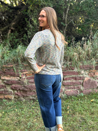 A woman wearing a multicolor knit long-sleeve top and blue jeans stands outdoors in front of a stone wall and greenery, looking back over her shoulder.