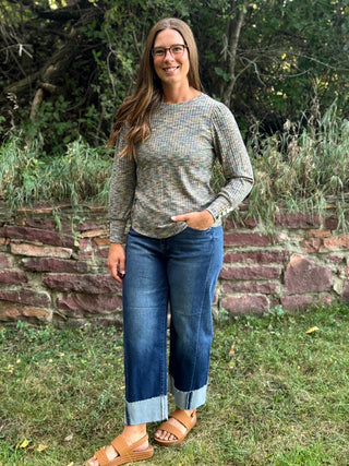 A woman wearing a long-sleeve knit sweater with a multicolor flecked pattern, blue cuffed jeans, and tan sandals, standing outdoors in front of a stone wall and greenery.