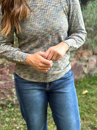 A person wearing a long-sleeve knit top with a multicolor heathered pattern in muted blue, beige, and green tones, paired with blue jeans, standing outdoors in front of a blurred stone wall and grass.