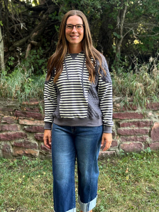 A woman wearing a black and white striped hoodie with gray knit side panels and blue jeans, standing outdoors in front of a stone wall and green trees.