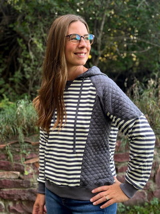 A woman wearing a mixed-media hoodie with navy and cream horizontal stripes on the body and sleeves, and dark gray quilted panels on the hood and shoulders, posing outdoors in front of a stone wall and green foliage.