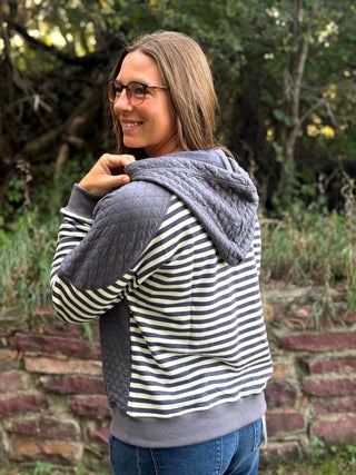 A woman wearing a gray quilted hooded sweatshirt with a navy and cream striped body, photographed outdoors in front of a brick wall and green foliage.