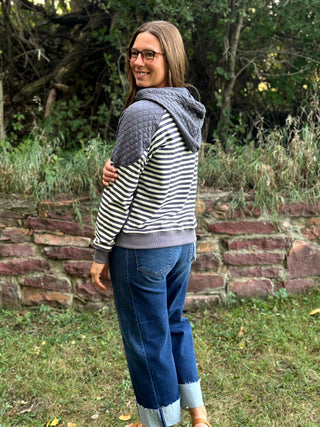A woman wearing a gray and black striped hoodie with a gray quilted shoulder panel, blue jeans, and brown boots, standing outdoors in front of a brick wall and greenery.