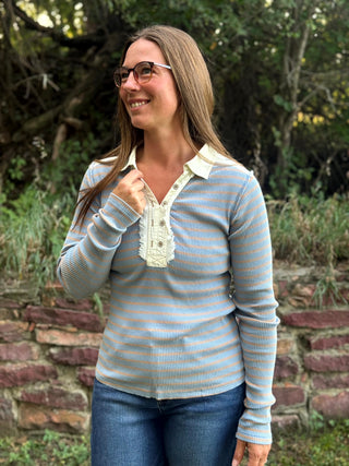 A woman wearing a light blue and white striped long-sleeve henley shirt with a cream collar and button placket, paired with blue jeans, standing outdoors in front of a stone wall and green foliage.