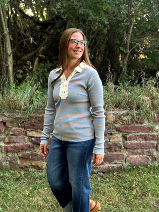 A woman wearing a light blue and gray striped long-sleeve shirt with a white lace-up collar, paired with blue jeans, standing outdoors in front of a stone wall and trees.