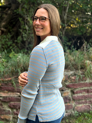 A woman with glasses and brown hair is shown from the back wearing a light blue and white striped ribbed long-sleeve polo shirt, standing outdoors in front of a stone wall and green foliage.