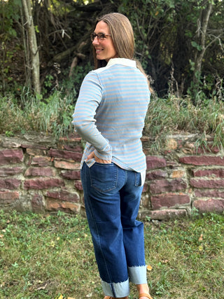 A woman wearing a light blue and white striped long-sleeve top and dark blue jeans stands outdoors in front of a stone wall and trees, photographed from the back.