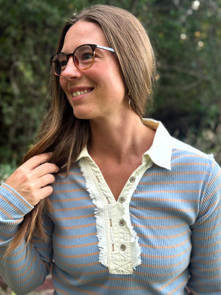 A woman with long brown hair and glasses wearing a light blue and orange striped long-sleeve top with a cream ruffled placket, photographed outdoors against a blurred green background.