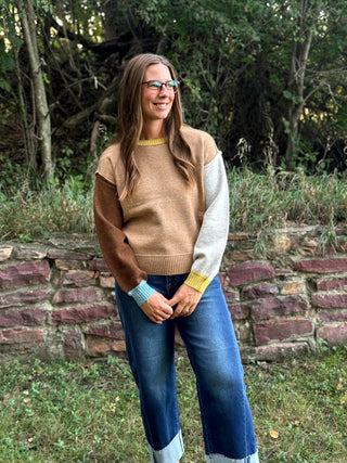 A woman wearing a color-block knit sweater with brown, tan, and light gray sections, blue jeans, and glasses, standing outdoors in front of a stone wall and trees.