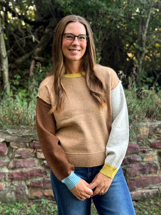 A woman wearing a color-block knit sweater with tan, brown, light gray, and light blue sections, standing outdoors in front of a stone wall and green foliage.