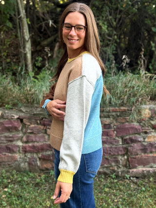 A woman wearing a color-blocked knit sweater with beige, light gray, light blue, and yellow panels, standing outdoors in front of a stone wall and greenery.