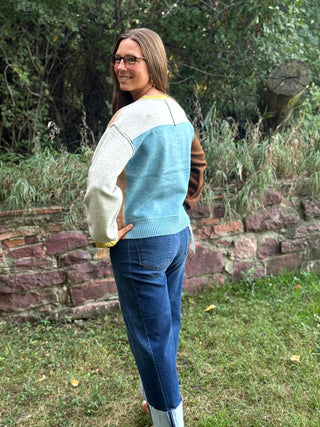 A woman wearing a color-block knit sweater with cream, light blue, and brown sections, paired with blue jeans, standing outdoors in front of a stone wall and green foliage.