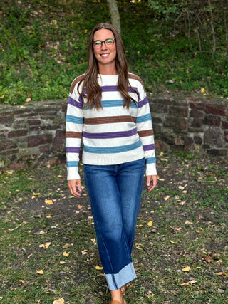A woman wearing a white knit sweater with horizontal stripes in purple, brown, and teal, paired with blue jeans, standing outdoors in front of a stone wall and green foliage.