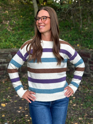 A woman wearing a cream knit sweater with horizontal stripes in purple, blue, and brown, paired with blue jeans, standing outdoors in front of a blurred green background.