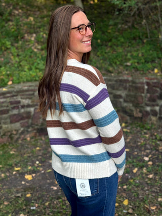 A woman wearing a cream knit sweater with brown, purple, and blue horizontal stripes, paired with blue jeans, photographed outdoors in a park setting.