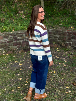 A woman wearing a striped knit sweater, blue jeans, and brown sandals stands outdoors in a grassy area with a stone wall behind her, looking over her shoulder.