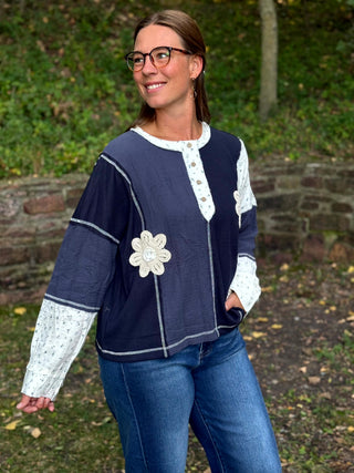 A woman wearing a navy blue long-sleeve top with white lace trim and a white floral applique, paired with blue jeans, standing outdoors in front of a stone wall and green foliage.