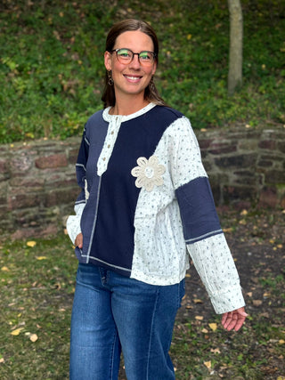 A woman wearing a patchwork knit cardigan with navy and white sections, a large white floral applique on the chest, and blue jeans, standing outdoors in front of a stone wall and green foliage.