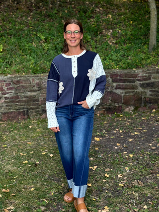 A woman wearing a navy blue and white patchwork long-sleeve top with floral embroidery, blue jeans, and brown sandals, standing outdoors in front of a stone wall and green foliage.