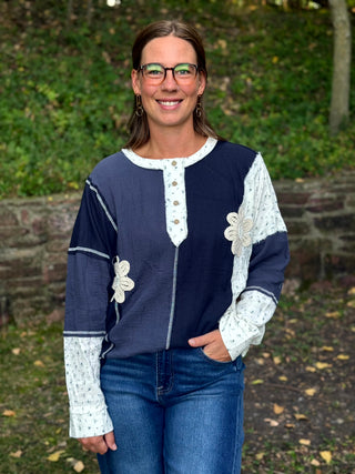 A woman wearing a navy blue and white patchwork long-sleeve top with floral applique and button details, paired with blue jeans, standing outdoors in front of a blurred green garden background.