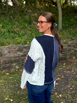 A woman wearing a patchwork knit sweater with navy blue and white sections, standing outdoors in a park-like setting with trees and a stone wall in the background.